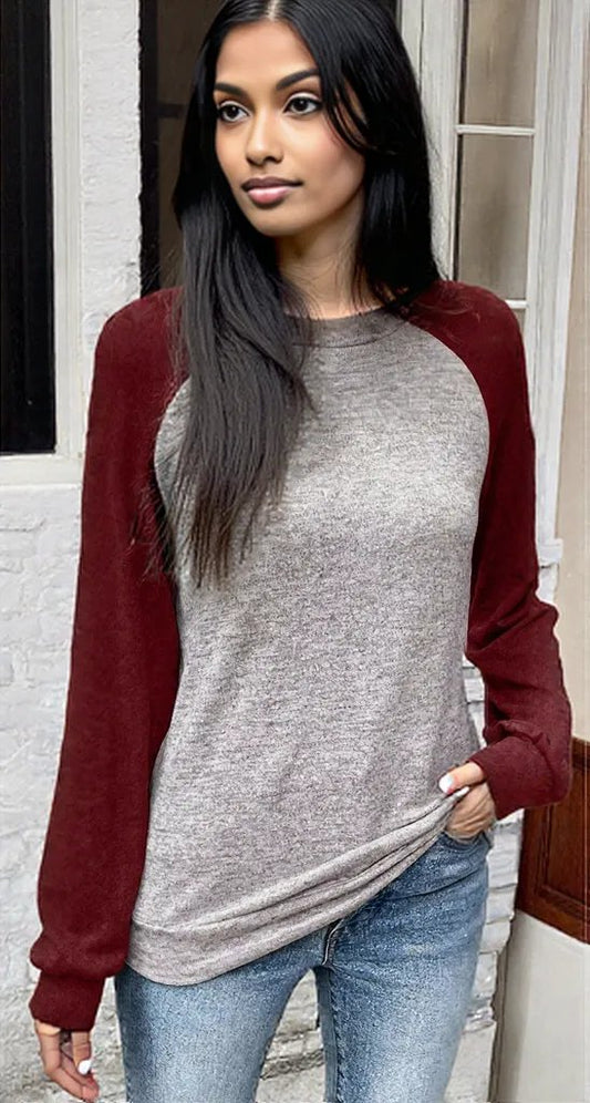 Woman wearing a gray and red long-sleeve shirt with blue jeans standing against a white wall.