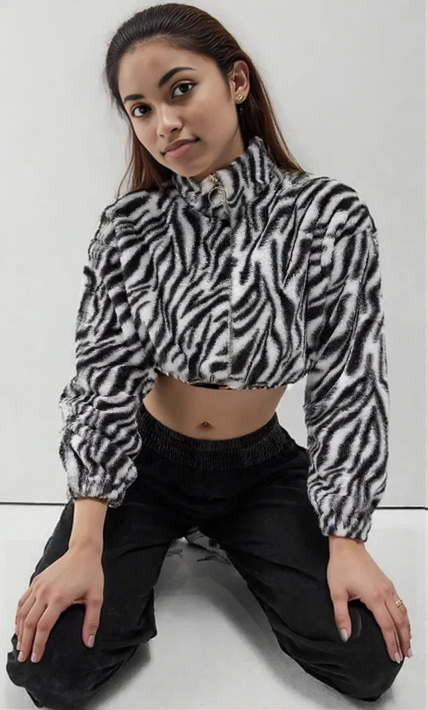 A young woman with long, straight brown hair with a center part poses against a plain white background. She is wearing a fuzzy, black-and-white zebra-print cropped zip-up hoodie and low-rise dark gray sweatpants, revealing her midriff. Her right hand, featuring a light-colored manicure, rests on her hip.