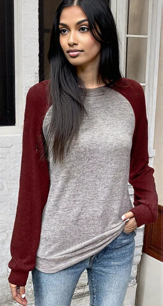 Woman wearing a gray and red long-sleeve shirt with blue jeans standing against a white wall.